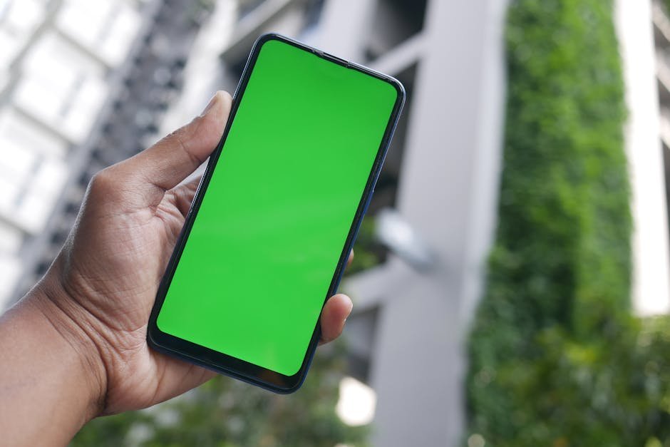 Hand holding a smartphone with a green screen outdoors, suitable for mockup or advertising.