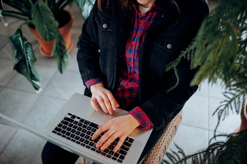 Close-up of hands typing on a laptop amidst houseplants, wearing a denim jacket and plaid shirt.