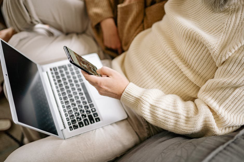 Adult in a cozy sweater multitasking with a smartphone and laptop indoors.